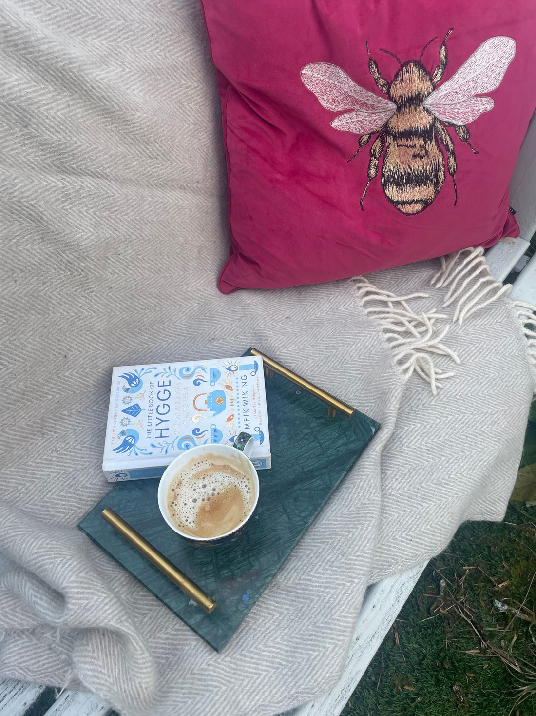 This picture shows a dark green tray with a cup of coffee and book. There's a grey blanket and a red cushion with a bee on it.