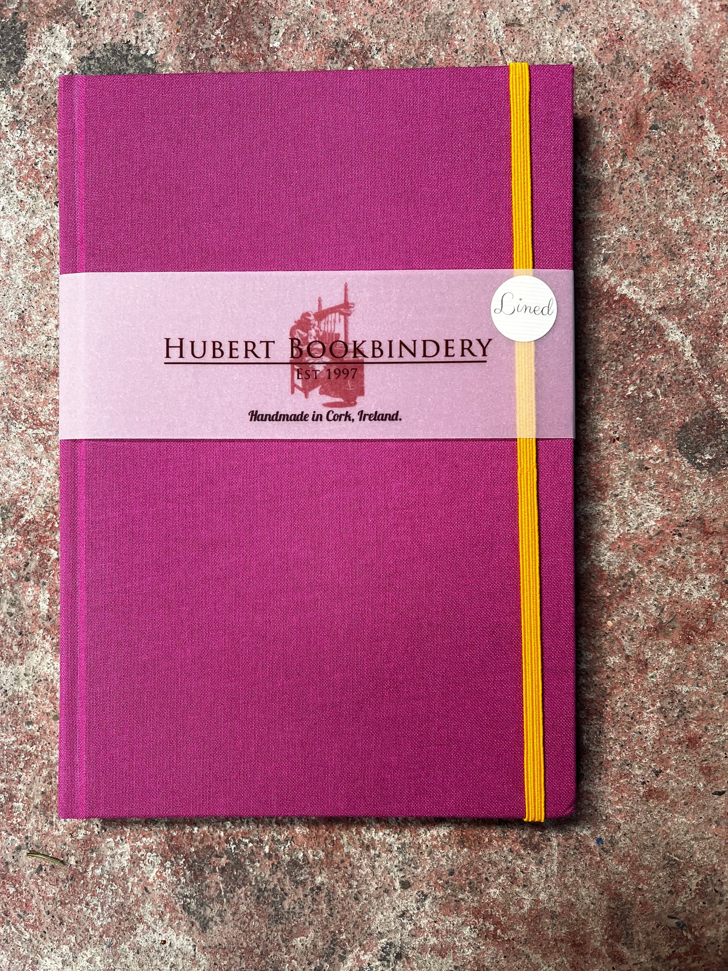 Purple notebook with a yellow strap on a textured surface, featuring 'Hubert Bookbindery' branding.