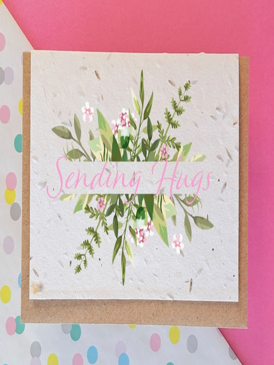This image shows a card called sending hugs. The card is made out of little seeds and can be planted. There is a picture of flowers on the front and the writing is in pink.