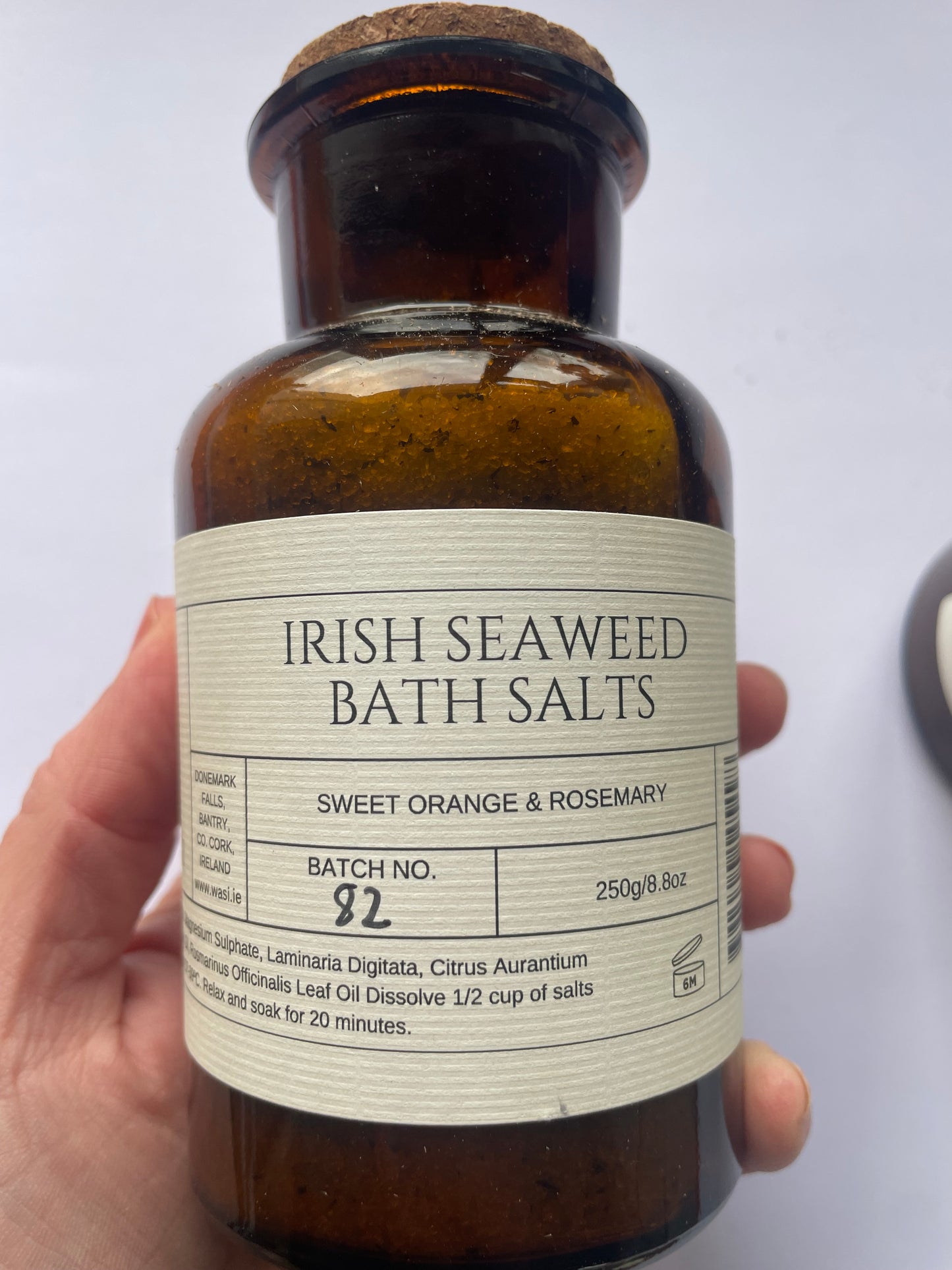 This picture shows a hand holding a brown bottle with a cream label that says 'Irish Seaweed bath salts' they are sweet orange and rosemary. It is an 8 oz bottle or 250g.