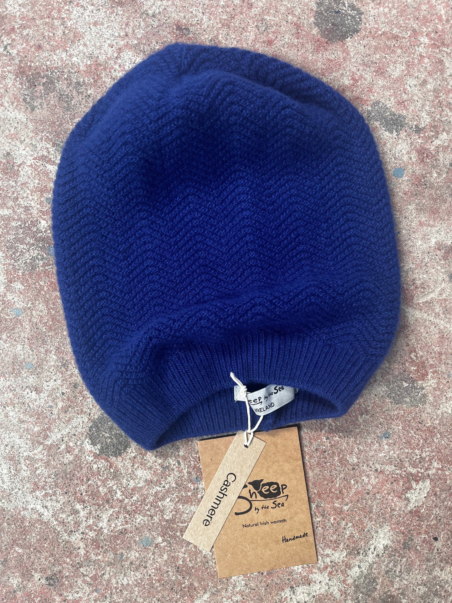 Blue knitted beanie on a textured surface with a brand tag.