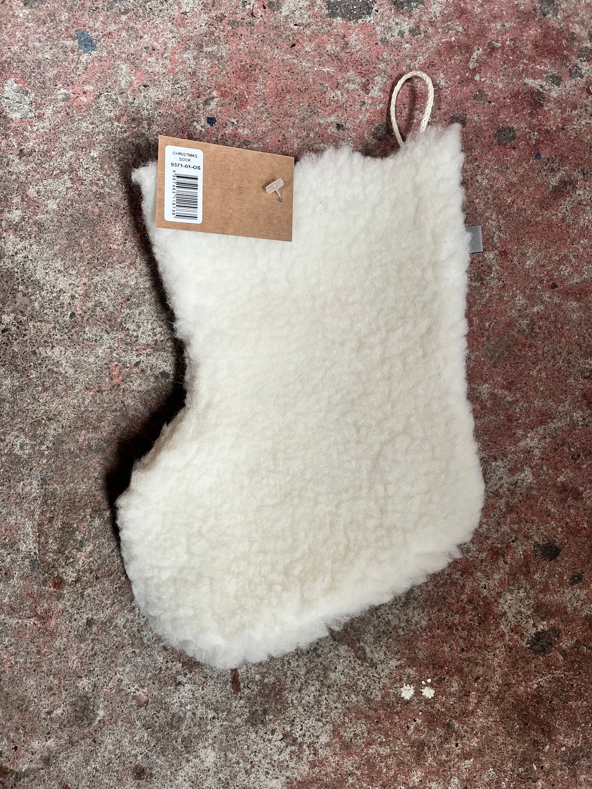 A fluffy off-white sheepskin or faux-sheepskin stocking with a brown paper hangtag and loop, placed on a textured concrete floor.