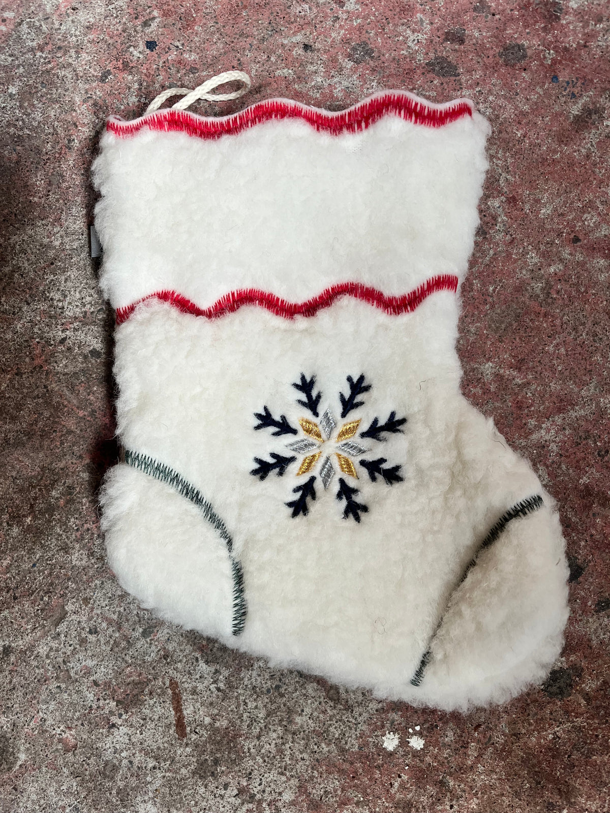 A white sheepskin mitten with a central embroidered snowflake design in black, gold, and white, featuring red and green stitched accents, laid flat on a textured brown surface.