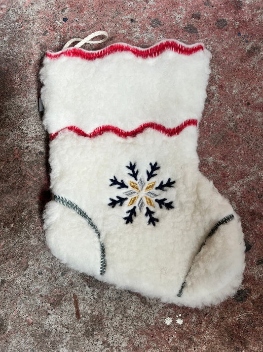 A white sheepskin mitten with a central embroidered snowflake design in black, gold, and white, featuring red and green stitched accents, laid flat on a textured brown surface.