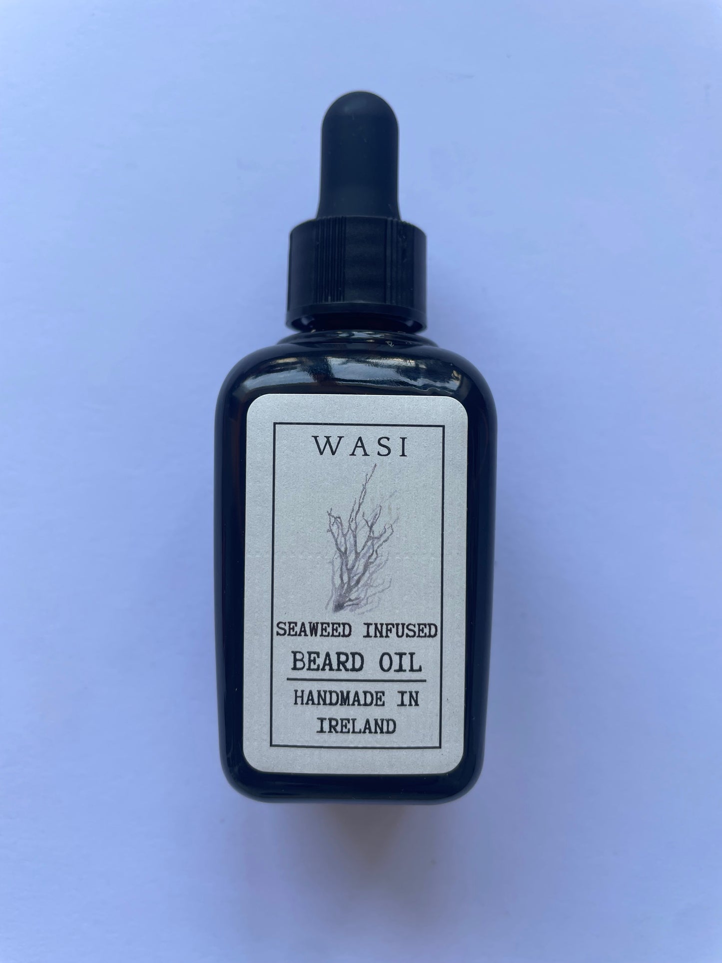 This image depicts a brown bottle with a black cork, it is a small bottle and on the front there is a cream label. It has the words 'seaweed infused beard oil, handmade in Ireland' written on the front as well as the brand logo WASI and a beautiful seaweed logo. 