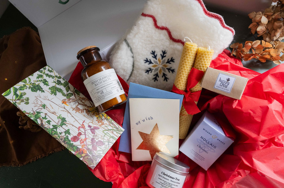 A curated gift basket or hamper filled with assorted self-care and home items, including a white knit stocking with a snowflake design, a brown glass bottle, a patterned card, a blue card, two yellow candle-like bars, a bar of soap, and a small jar, arranged on red tissue paper.