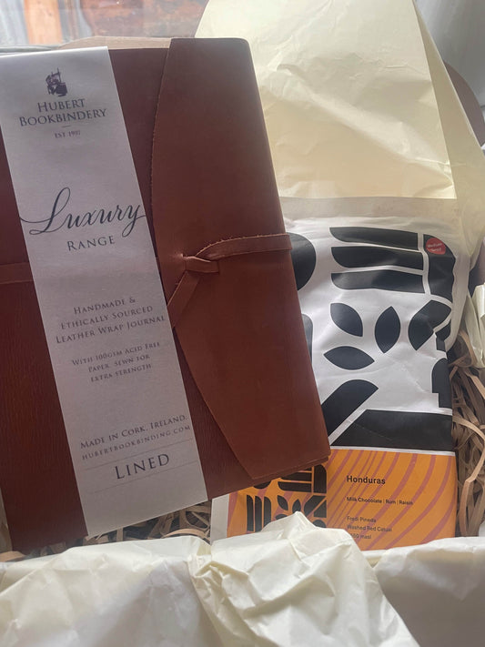 This is a picture of an opened gift box with tissue paper inside. It has a luxury leather bound journal and a bag of ground coffee. 