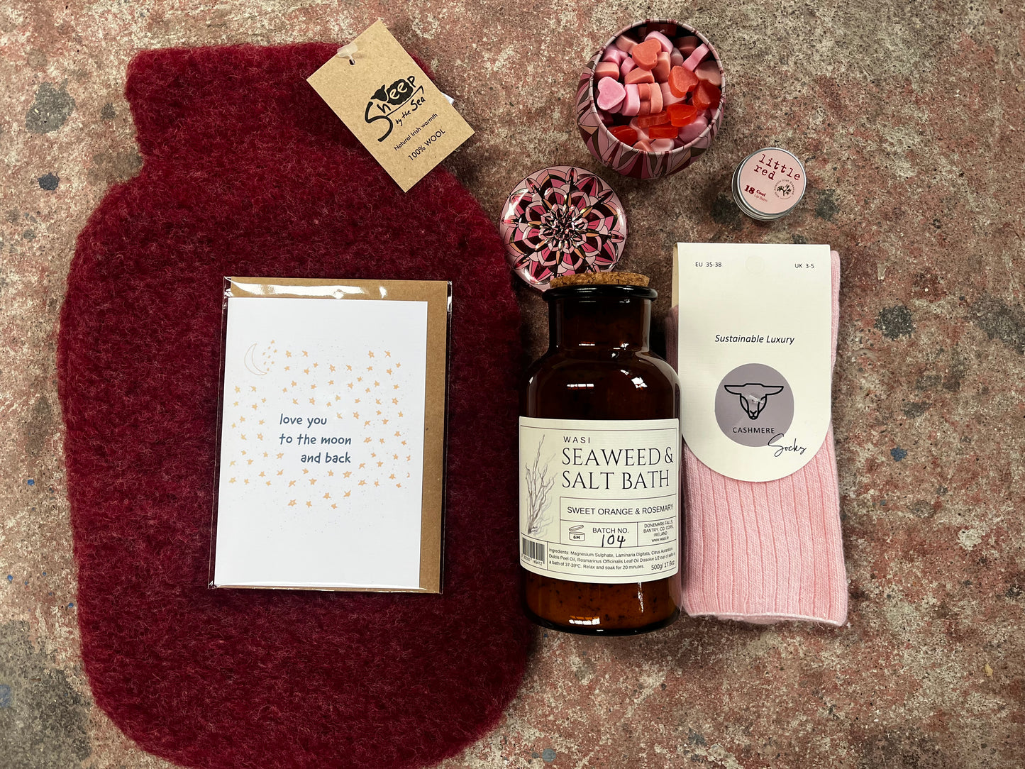 Collection of self-care items including a hot water bottle, bath salts, and a sock on a textured surface.