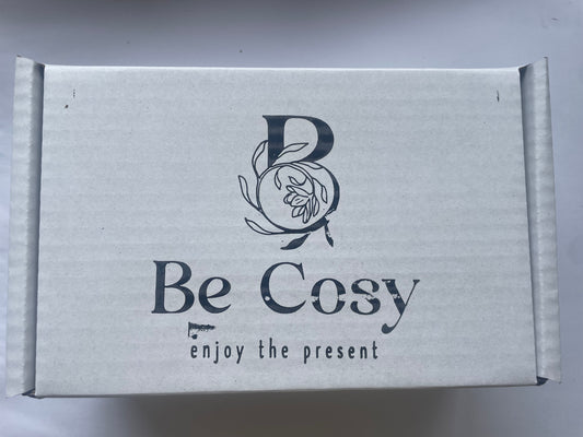 This is a picture of a  small white box with the words 'Be Cosy' enjoy the present n the front. There is a large B on top of the words Be Cosy with a hand drawn logo with jasmine flowers coming out of it. 
