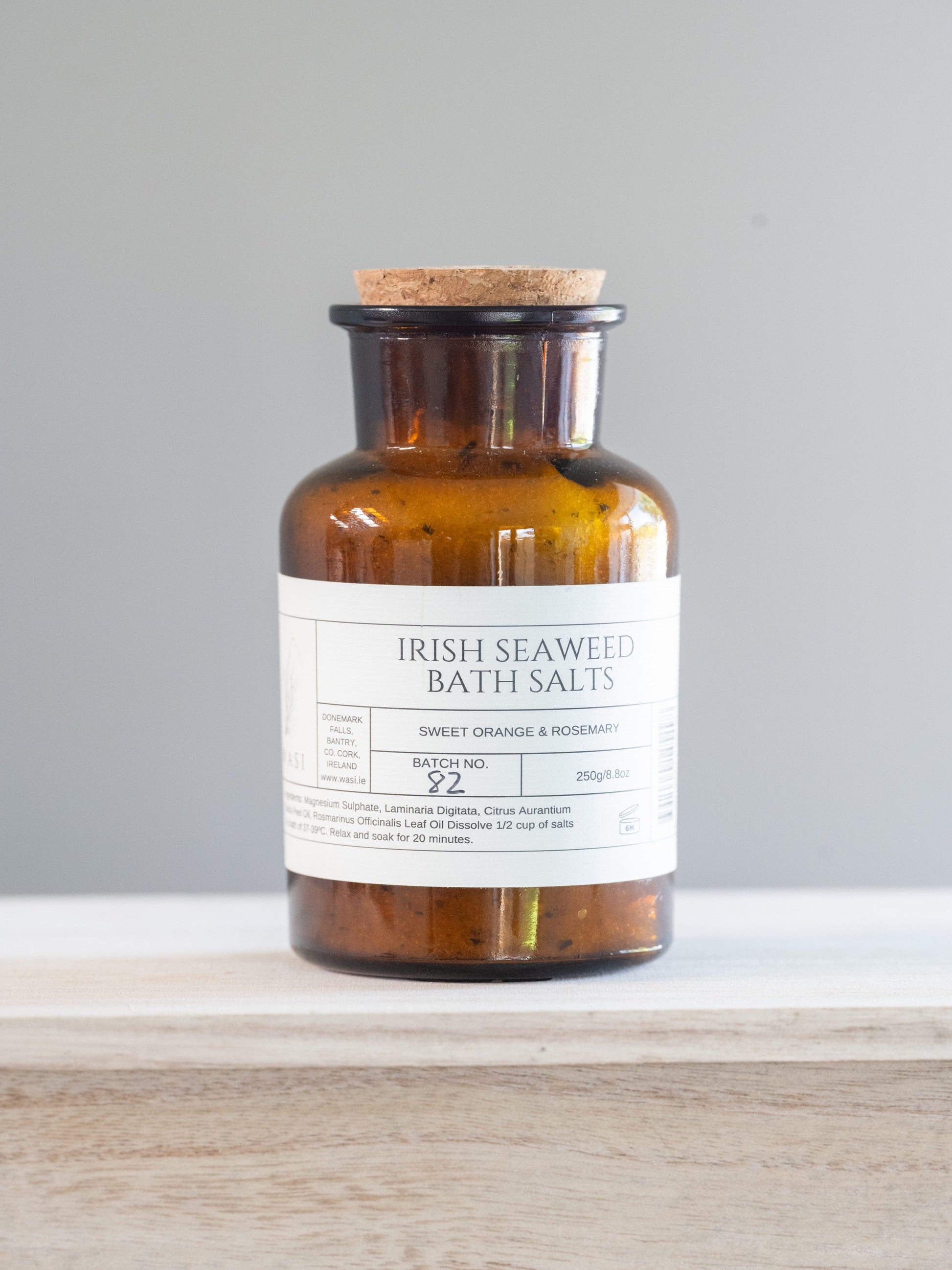 Bottle of Irish Seaweed Bath Salts on a wooden surface with a grey background. These are sweet orange and rosemary. 