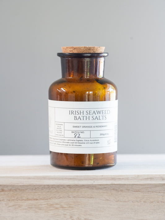 Bottle of Irish Seaweed Bath Salts on a wooden surface with a grey background. These are sweet orange and rosemary. 