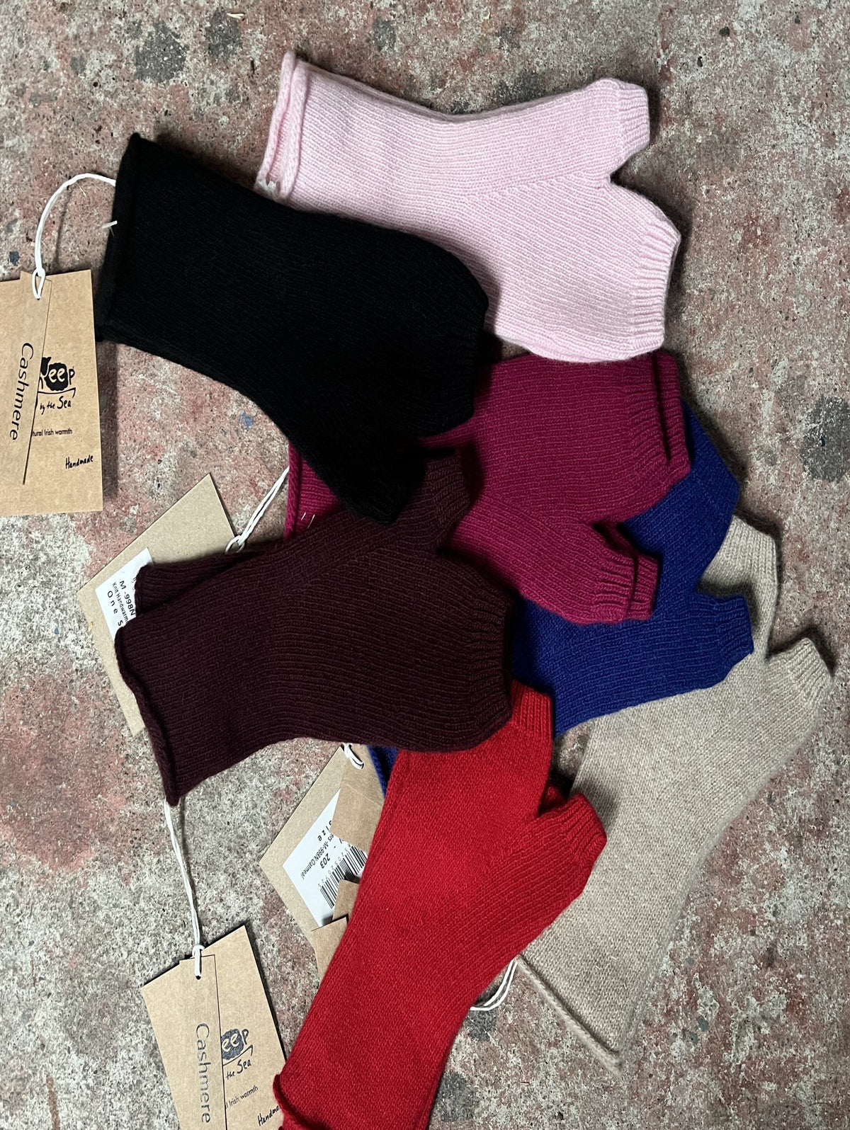 A pile of ribbed knit fingerless gloves in multiple colours, including black, light pink, burgundy, maroon, blue, beige, and red, laid out on a textured stone surface. Each sock has a paper hangtag attached.