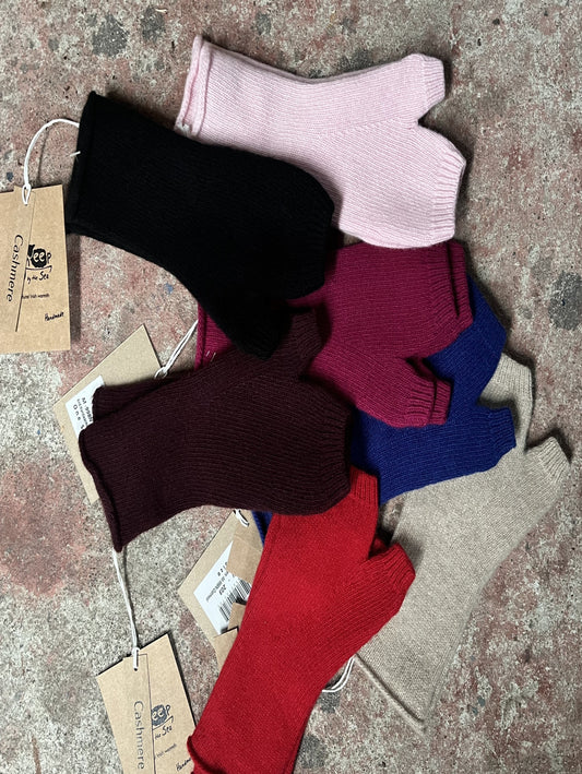 A pile of ribbed knit fingerless gloves in multiple colours, including black, light pink, burgundy, maroon, blue, beige, and red, laid out on a textured stone surface. Each sock has a paper hangtag attached.