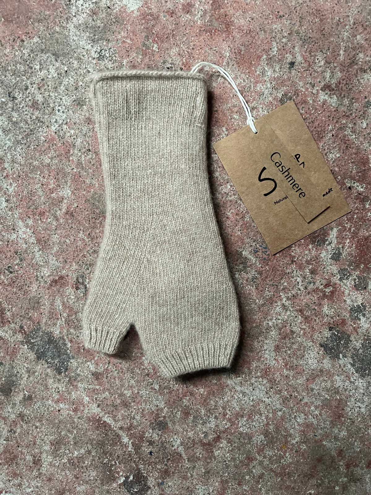 A single beige knit fingerless glove or mitten laid flat on a mottled stone surface, with a small kraft paper tag attached by a white string.