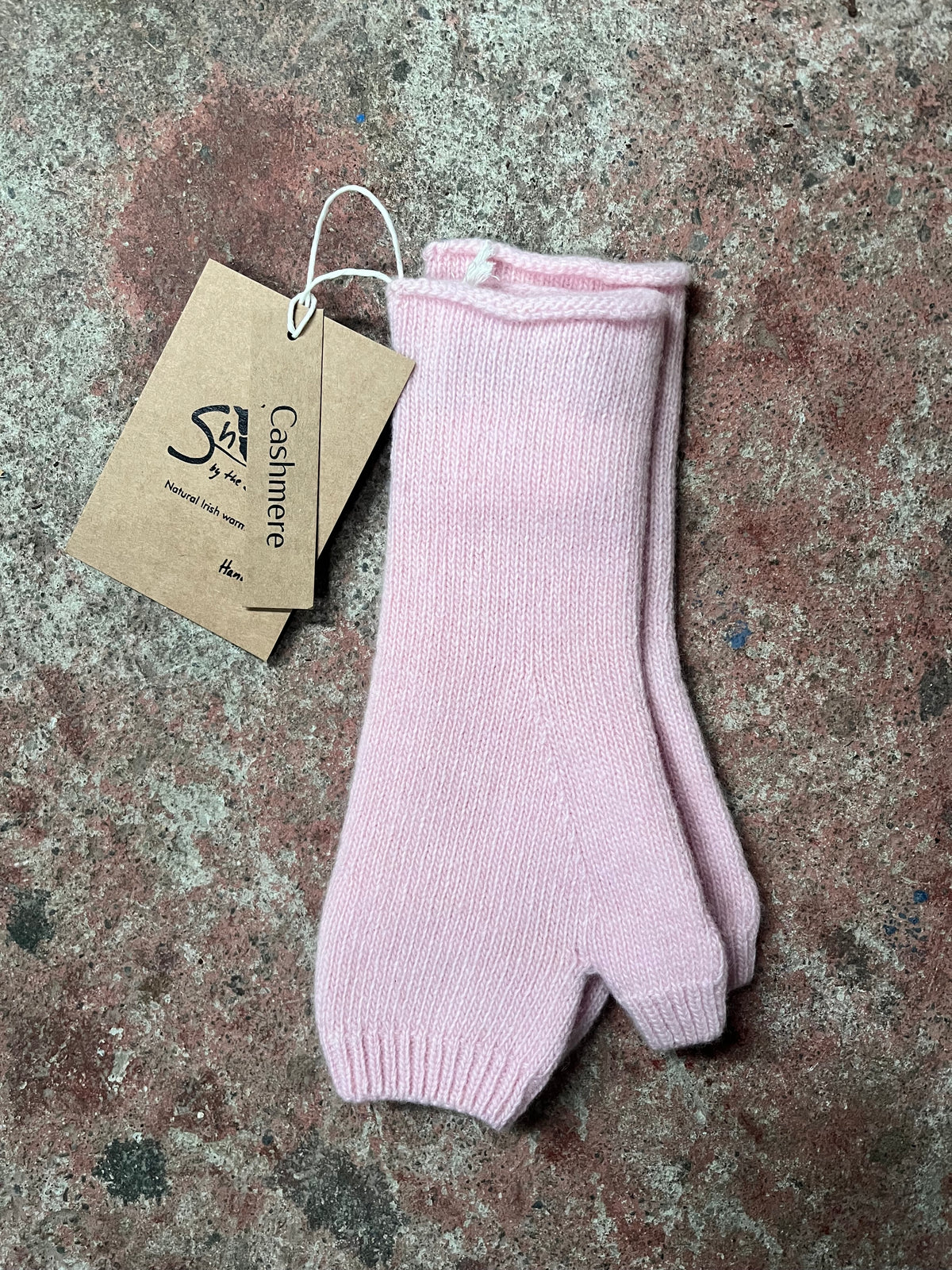 A pair of light pink ribbed knit split-toe booties with a brown paper tag reading 'Cashmere' and a stylized 'SA' logo, laid flat on a textured stone surface.