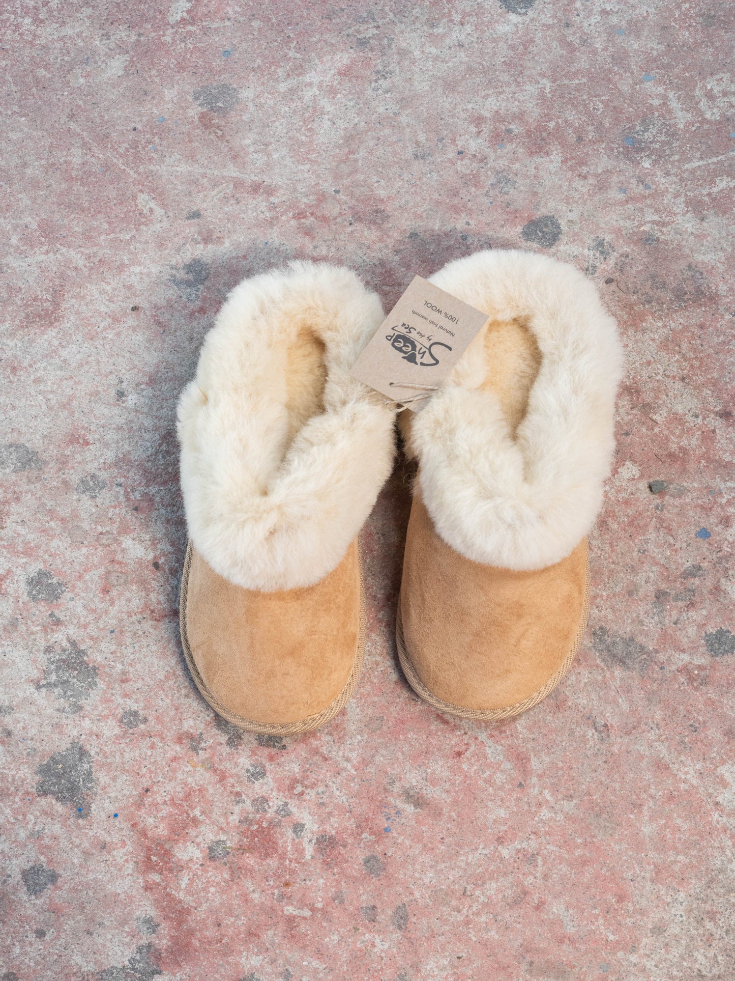 Pair of tan slippers with white fur lining on a textured pink surface. The slippers are 100% wool and made by sheep by the sea in Ireland.