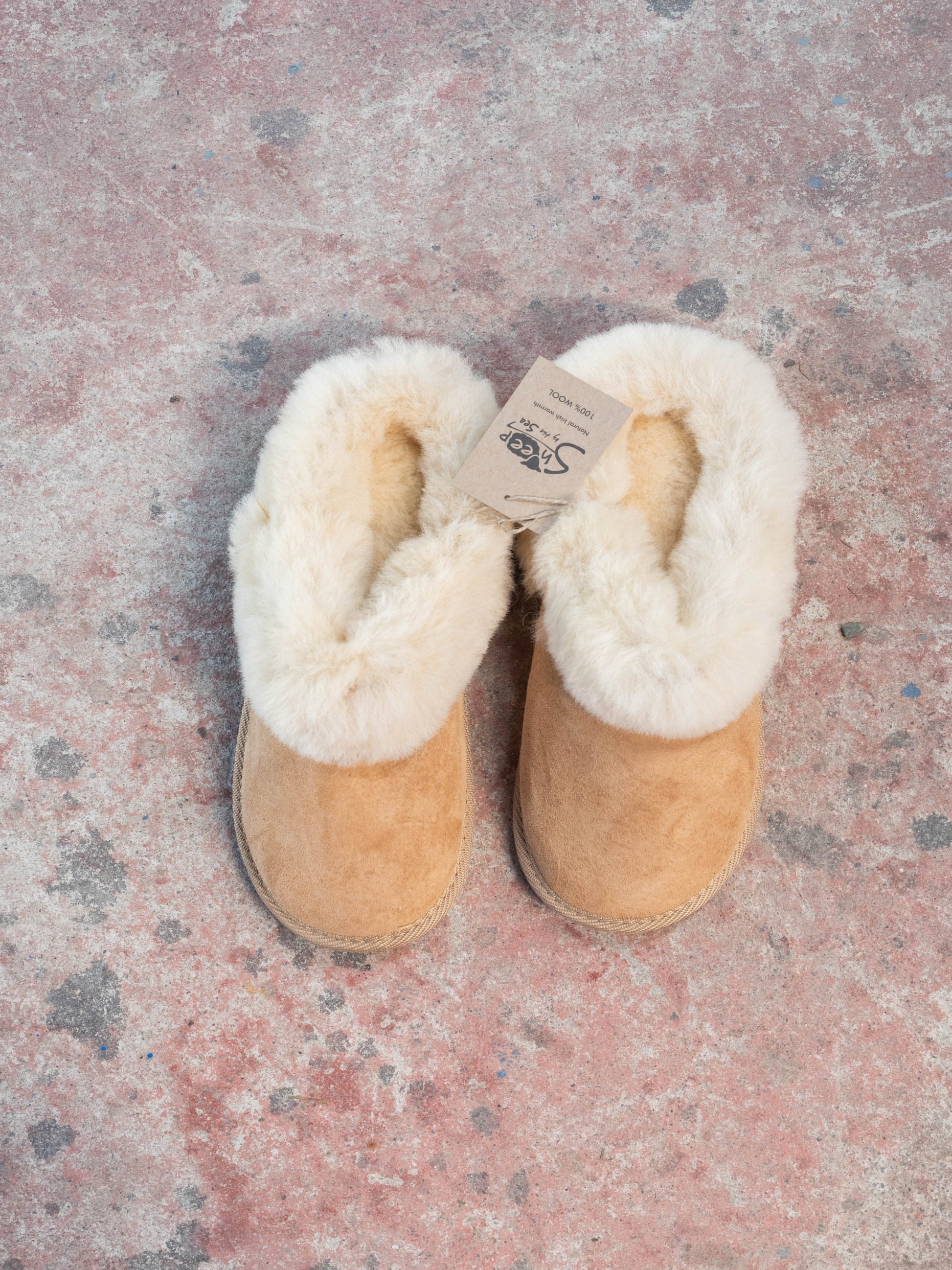Pair of tan slippers with white fur lining on a textured pink surface. The slippers are 100% wool and made by sheep by the sea in Ireland.
