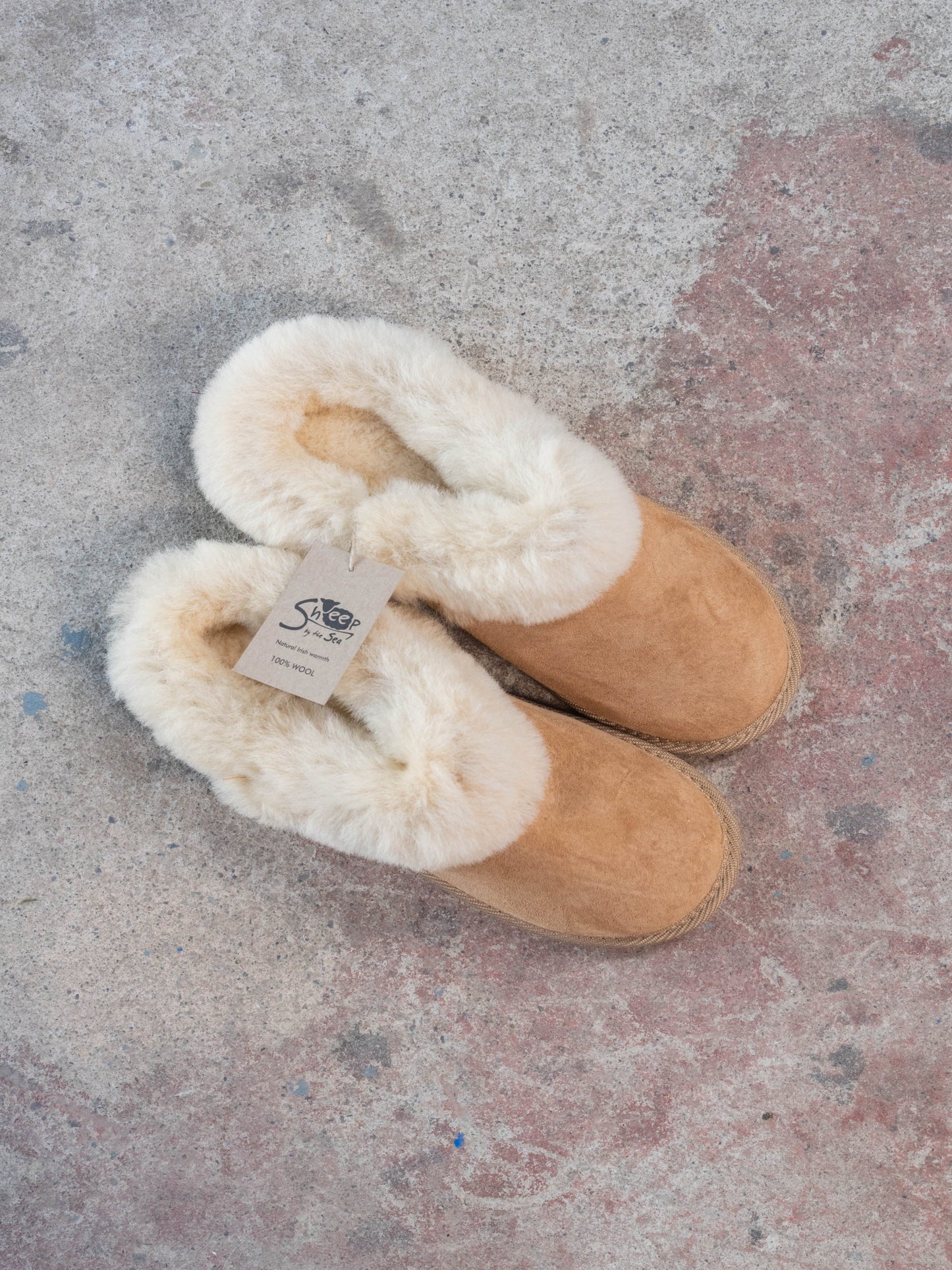 Pair of tan slippers with white fur lining on a textured surface. 100% wool inside. Made in Ireland by sheep by the sea.