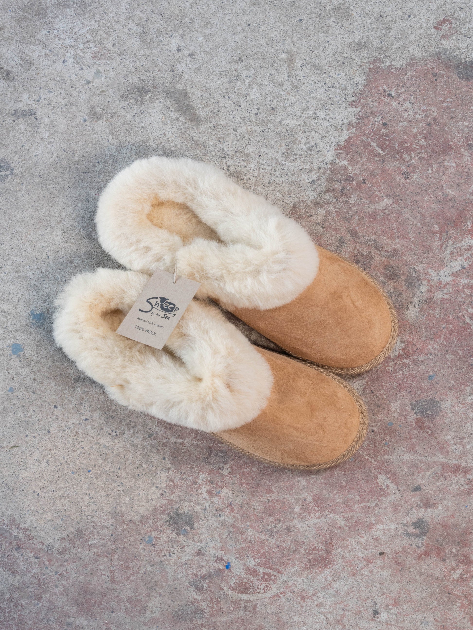 Pair of tan slippers with white fur lining on a textured surface. 100% wool inside. Made in Ireland by sheep by the sea.