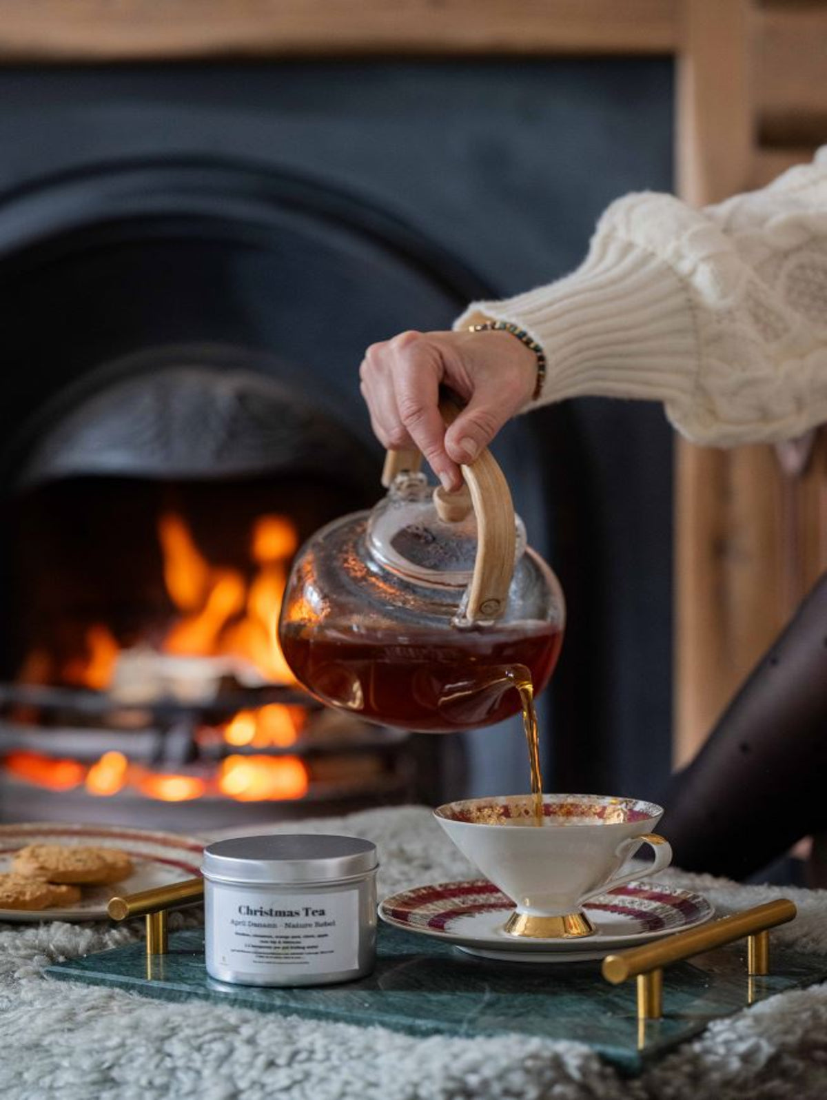 A person in a cream cable-knit sweater pours tea from a clear glass teapot into a white teacup with gold trim, set on a glass tray in front of a cozy fireplace. A silver tin labeled 'Christmas Tea' sits nearby.