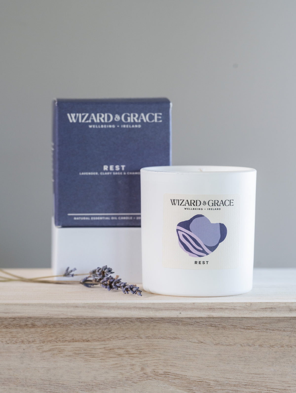 A white wax candle jar labeled 'WIZARD + GRACE' with a purple heart graphic and 'REST' scent, placed on a light wood surface in front of its navy and white product box. A small sprig of lavender lies beside the candle.