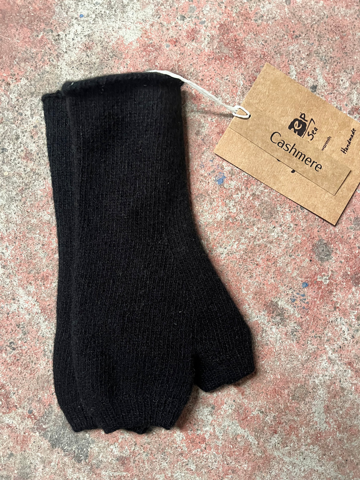 A single black ribbed fingerless glove or mitten laid flat on a mottled pinkish-beige surface, with a brown paper tag attached near the cuff. The tag shows the brand name Cashmere.