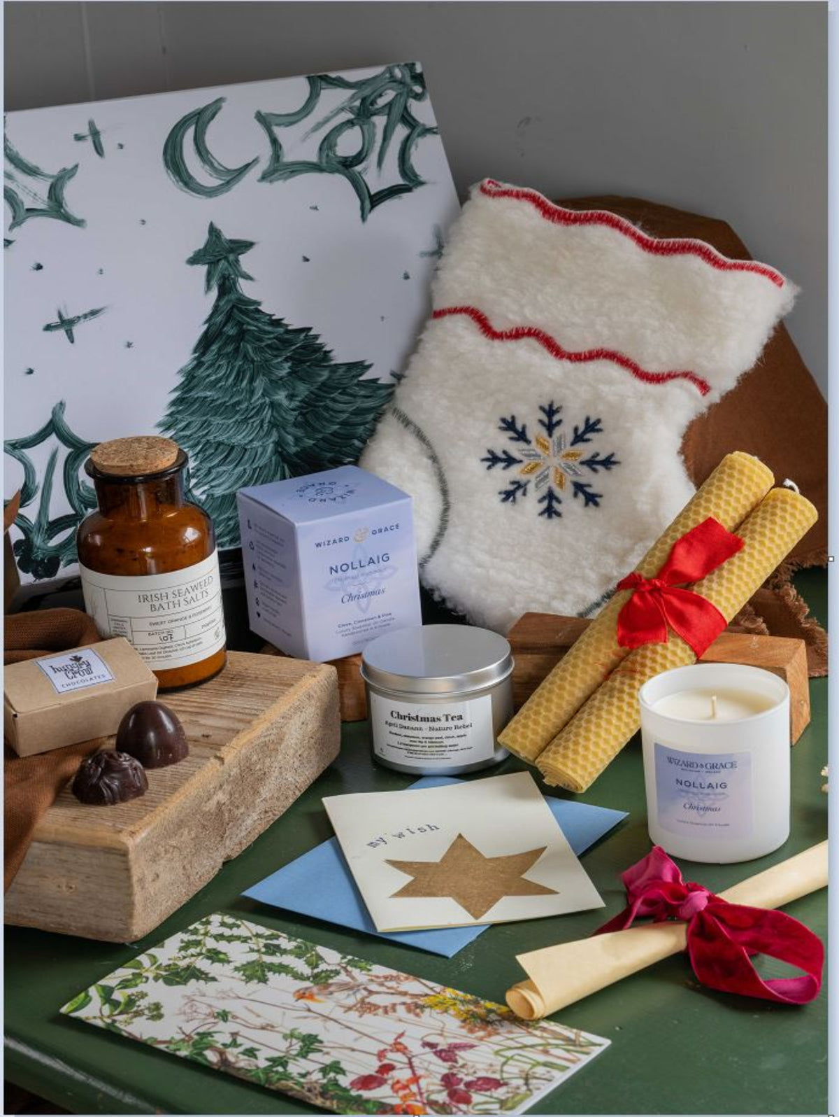 A styled gift set featuring a white Christmas stocking with a blue snowflake, a candle, a boxed tea, an amber bottle, beeswax candles, and a card set arranged on a green surface with a white Christmas tree print background.