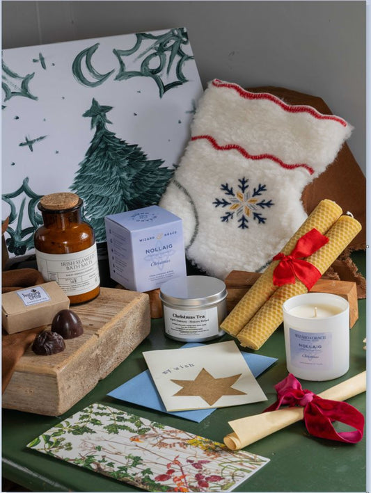 A styled gift set featuring a white Christmas stocking with a blue snowflake, a candle, a boxed tea, an amber bottle, beeswax candles, and a card set arranged on a green surface with a white Christmas tree print background.