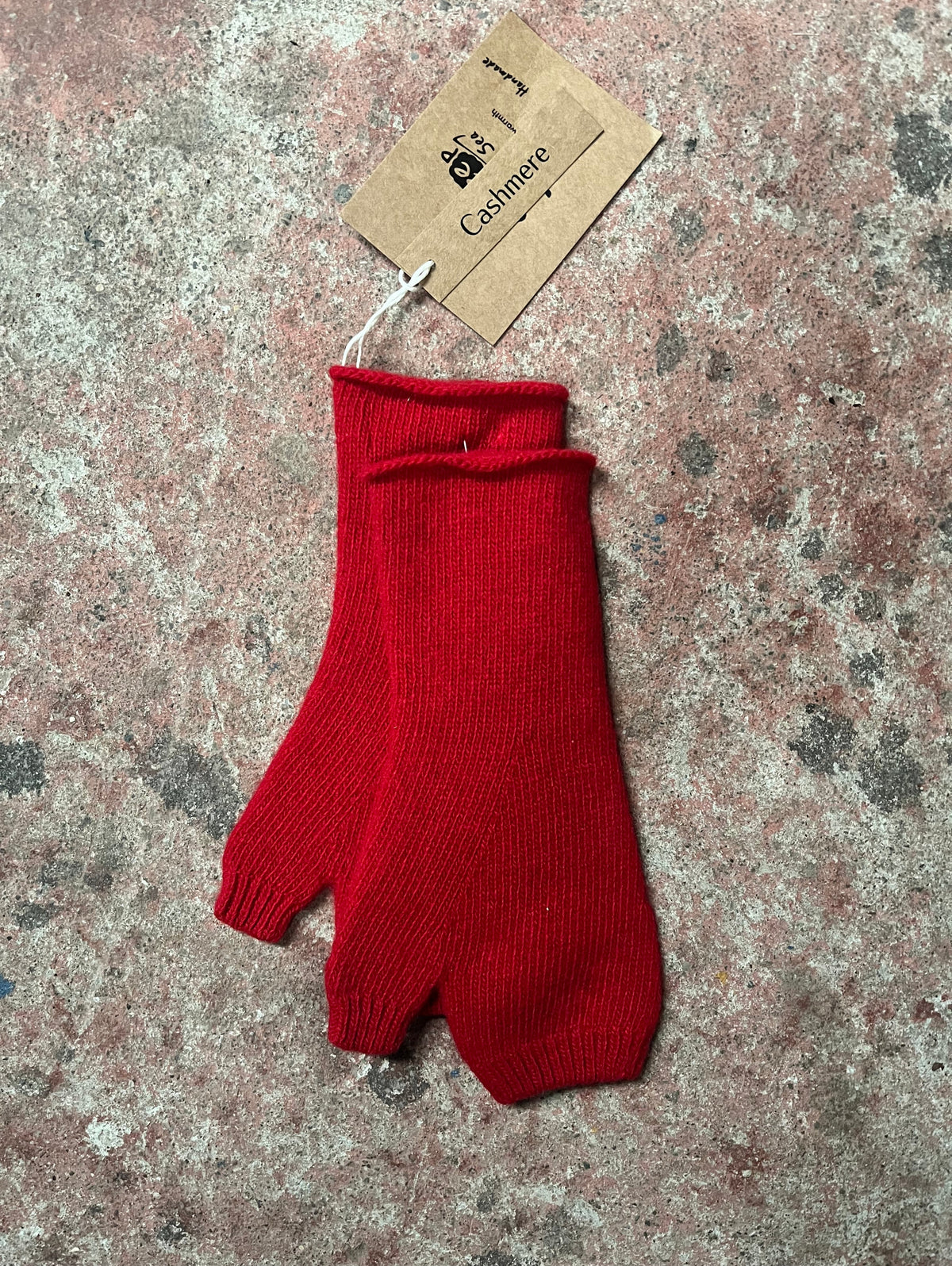 A single red fingerless knit glove with a brown paper tag attached, placed on a textured concrete surface.