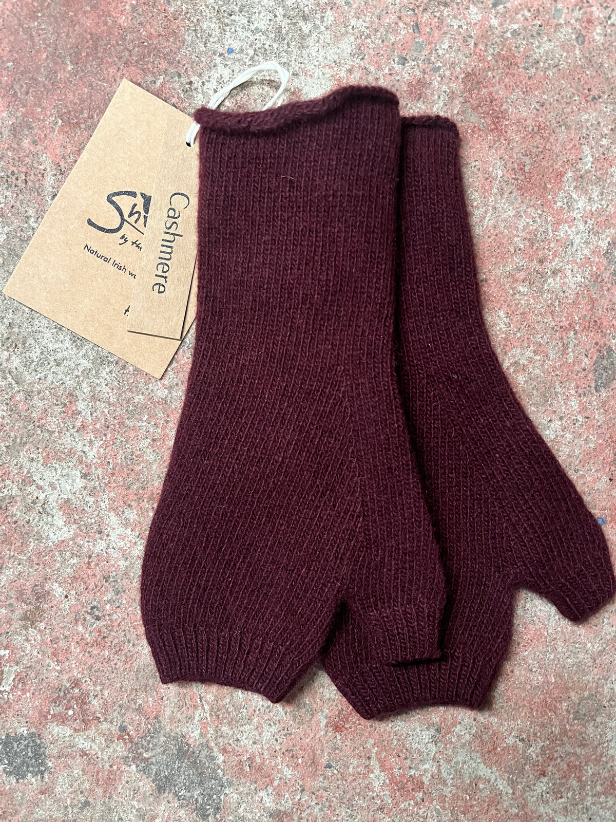 A pair of dark burgundy knit fingerless gloves or mittens with a brown paper tag that reads 'Cashmere', placed on a textured, speckled gray and pink surface.